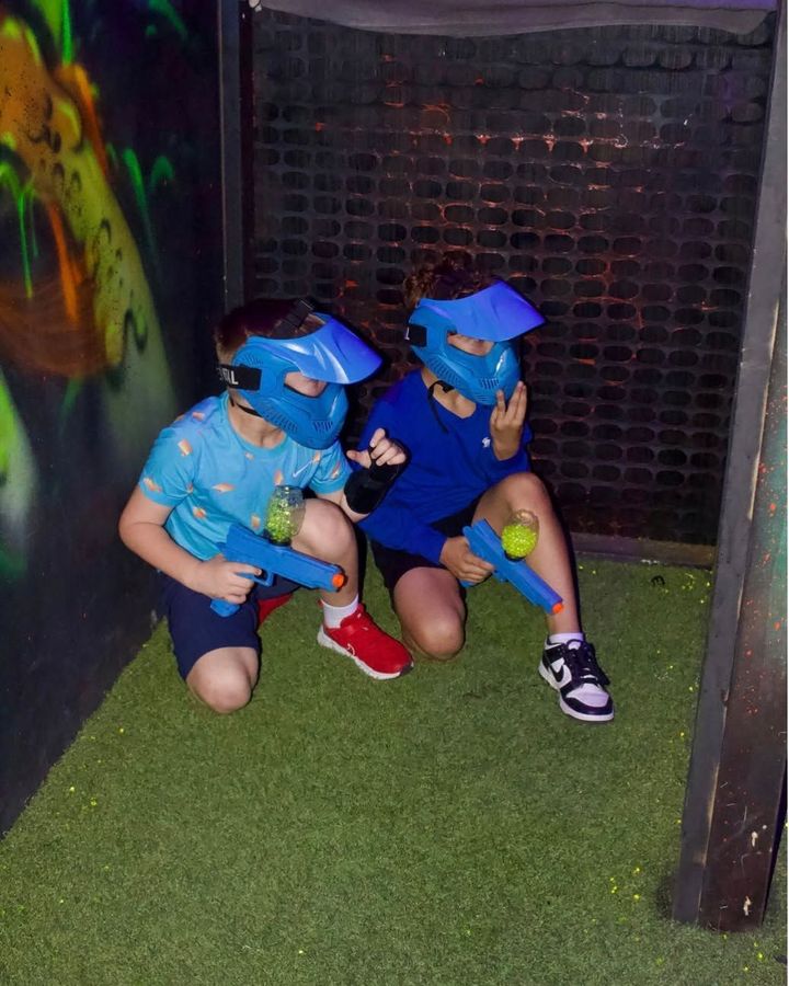 Two children in blue helmets crouch in a dark play maze, holding neon toy balls.
