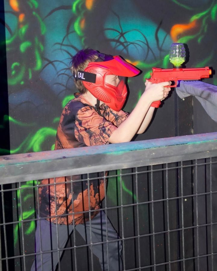 Person in a red visor aims an orange laser tag gun in a neon-lit arena.