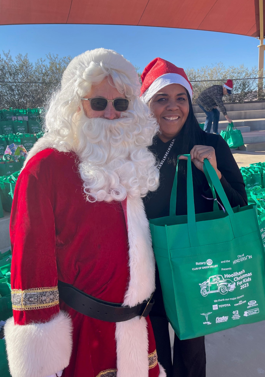 A man dressed as santa claus is standing next to a woman holding a green bag.