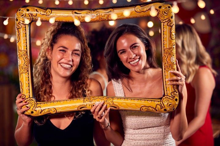Two women smiling and posing with a gold ornate frame at a festive event