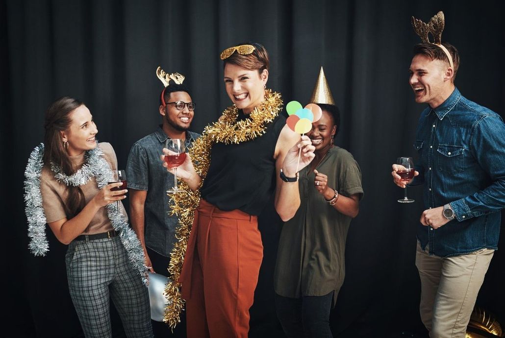 Group of friends celebrating at a party with drinks, wearing festive hats and garlands against a dark backdrop.