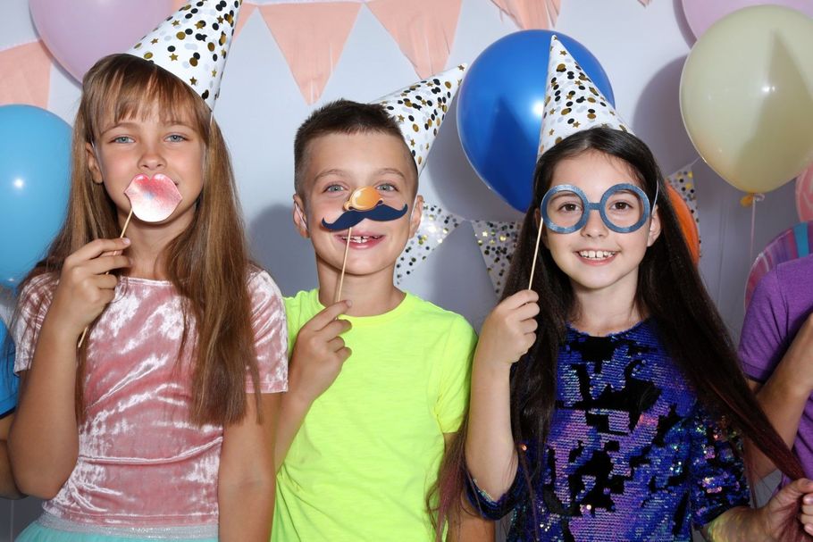 Three children at a birthday party wearing cone hats and holding playful props against pastel balloons.