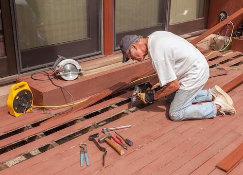 Worker kneeling on a wooden deck, using a power tool to repair broken boards, with tools nearby