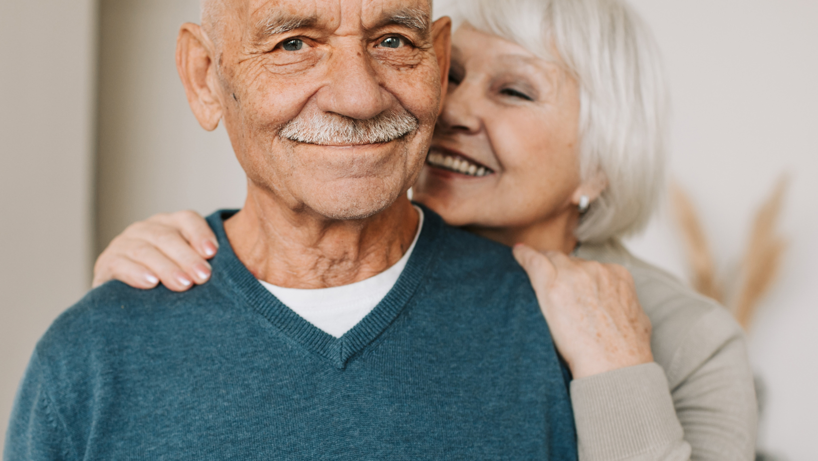 Smiling older couple embracing indoors, with one person in a teal sweater and the other in a light gray top