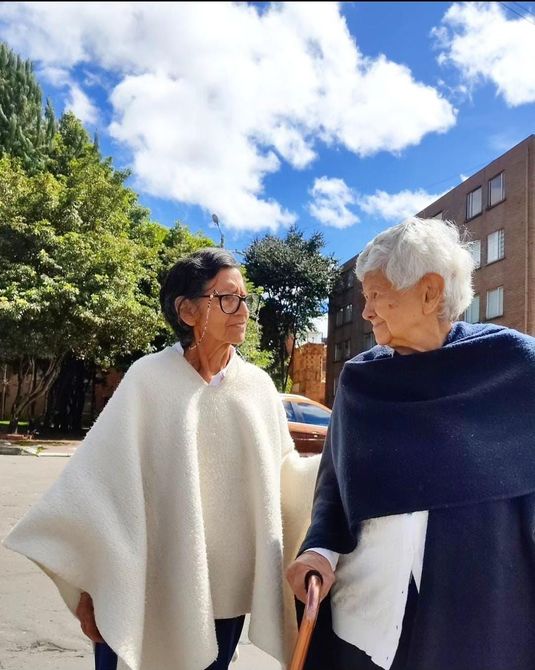 Dos personas con chales blancos y azul marino sonríen al aire libre en un soleado paseo del campus.