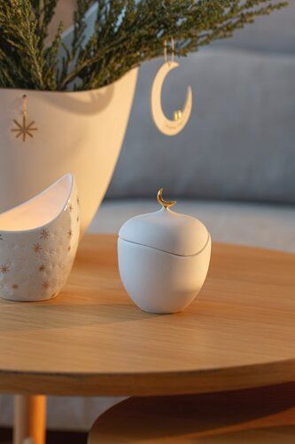 Een witte pot staat op een houten tafel naast een gespikkelde kop en vaas, met een ornament in de vorm van een halve maan.