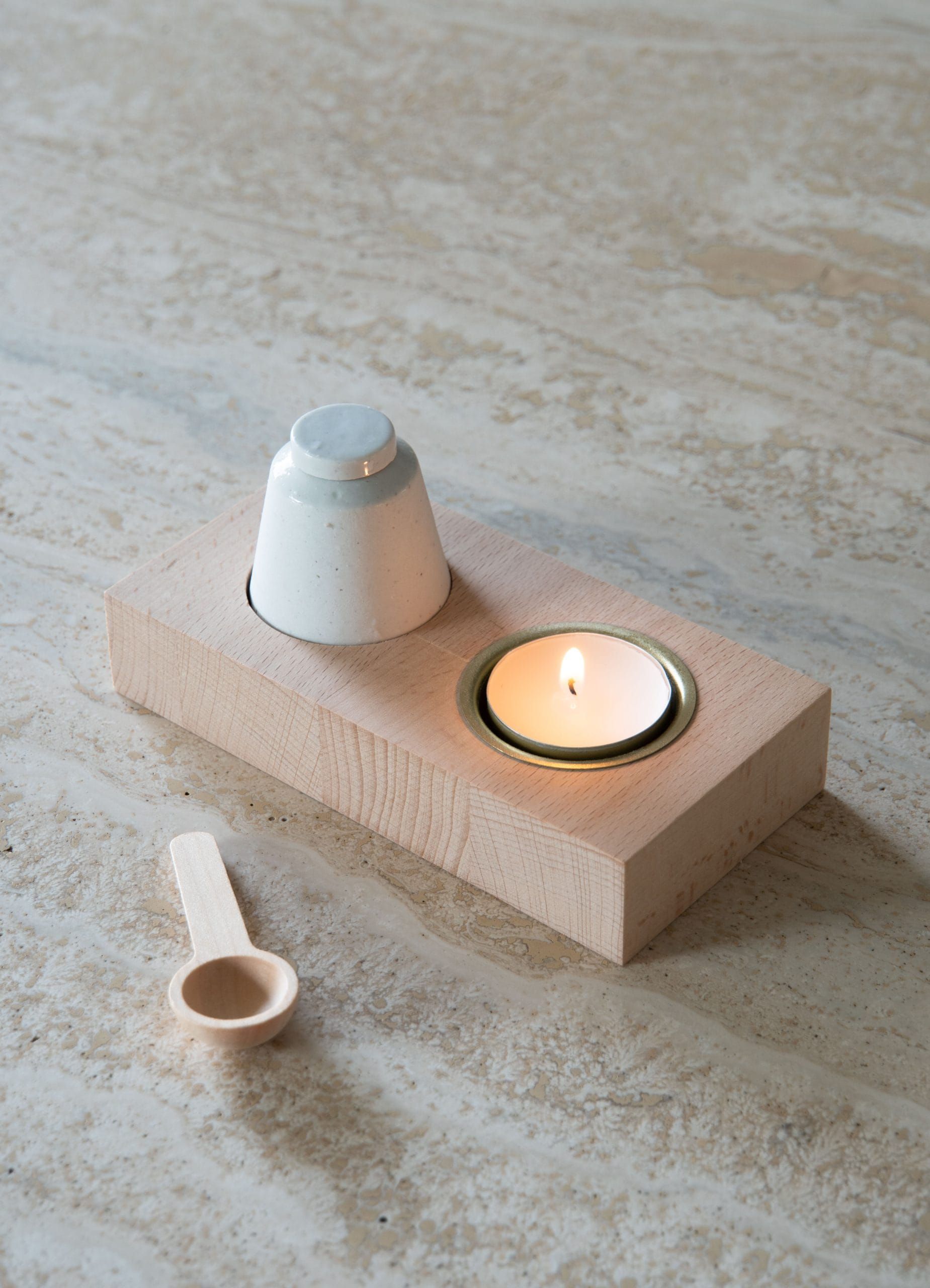 Kleine beige kaarsenhouder met een brandend theelichtje en een wit keramisch object op een lichtstenen ondergrond.