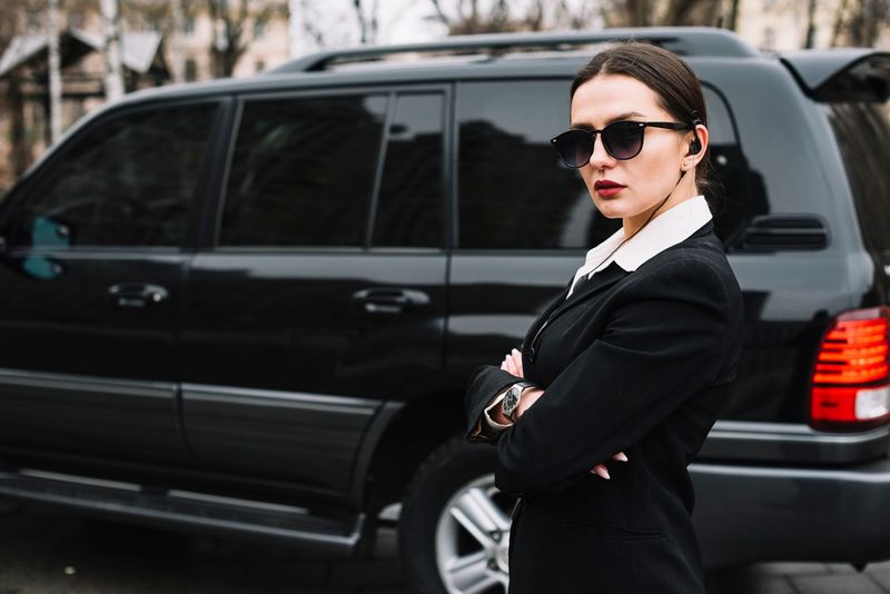 Woman in black suit and sunglasses standing beside a black SUV outdoors