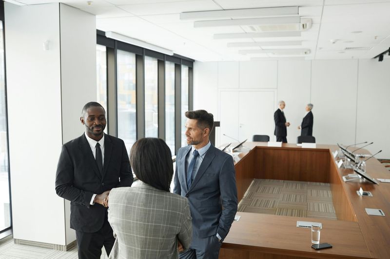 Three businesspeople in a bright conference room, shaking hands near a long table.