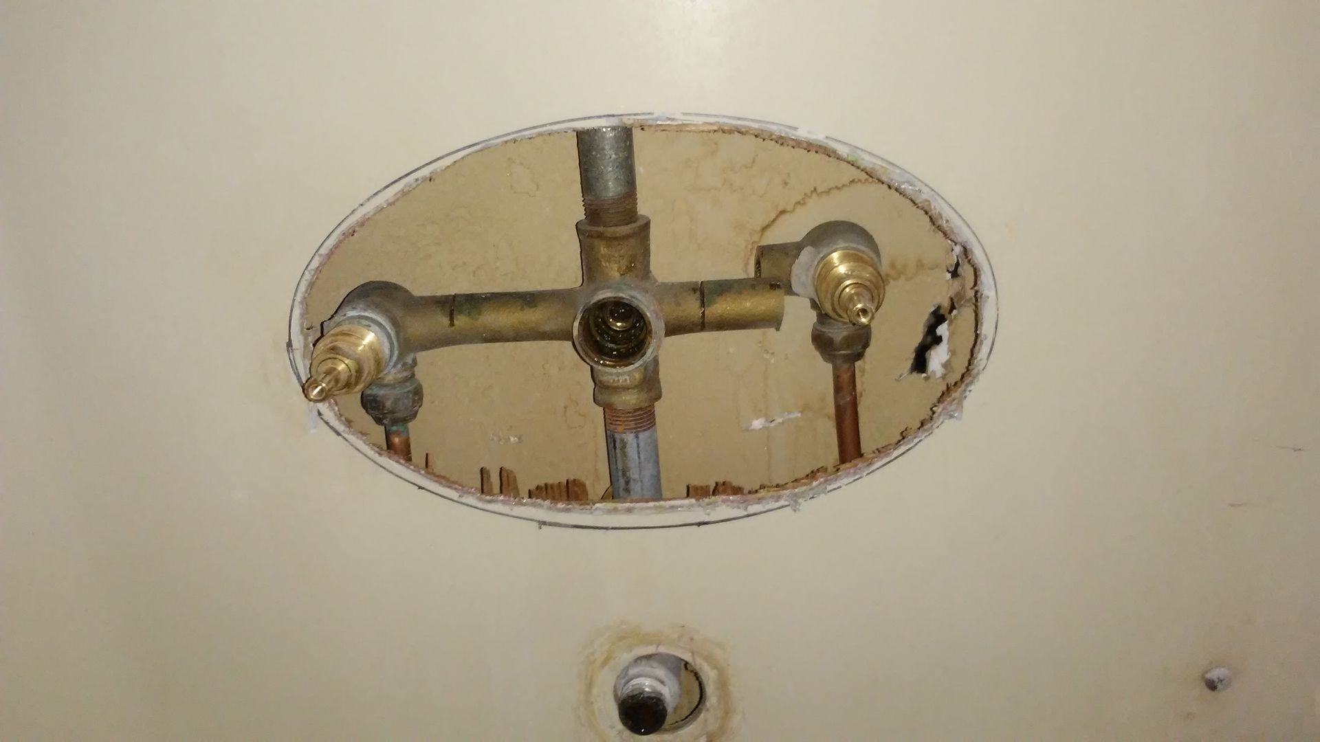 Older shower valve exposed through an oval opening, with separate hot and cold controls and threaded pipe sections.