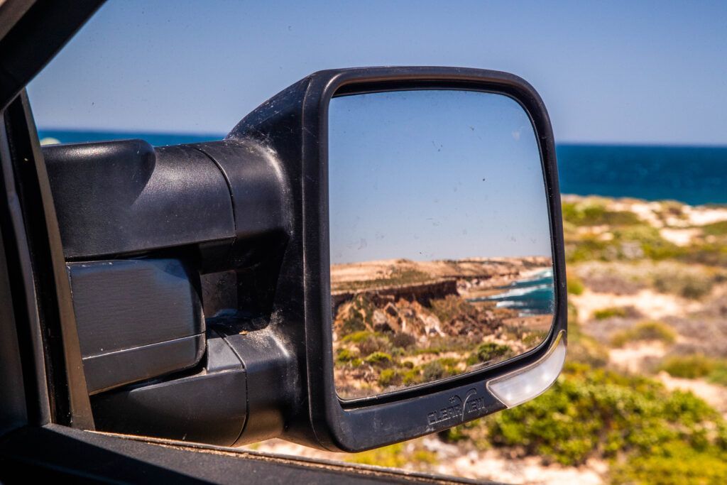 Car side mirror reflecting a sunny coastal road with cliffs and ocean beyond — Ol Mate 4x4 in Tuggerah, NSW