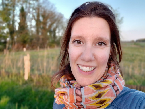 Een vrouw lacht in de buitenlucht, met een sjaal om. Op de achtergrond zijn een veld en bomen te zien.