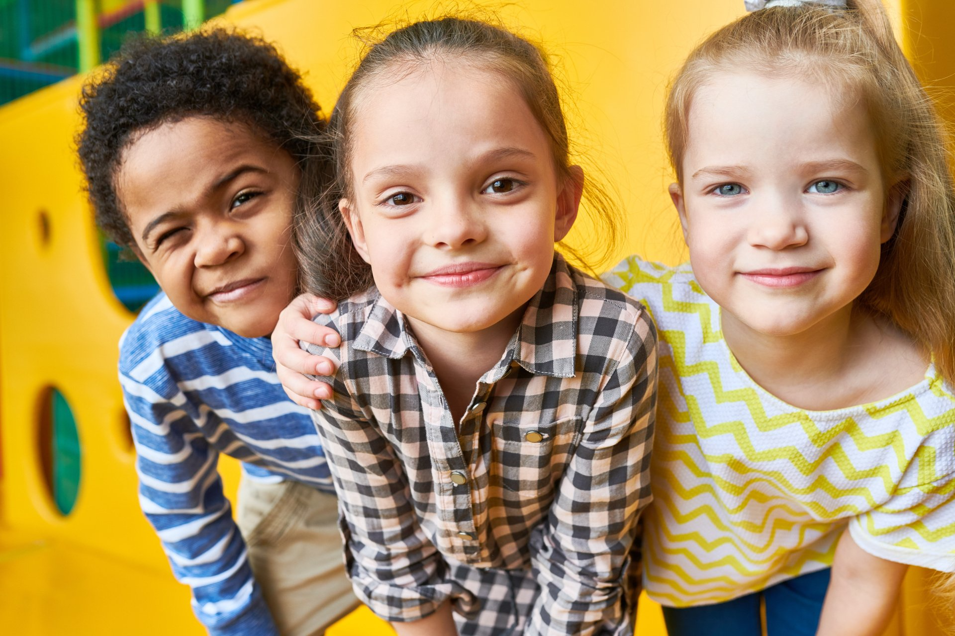 Drie kinderen lachen samen in een gele binnenspeeltuin. Een van de kinderen heeft zijn arm om een ander heen geslagen.