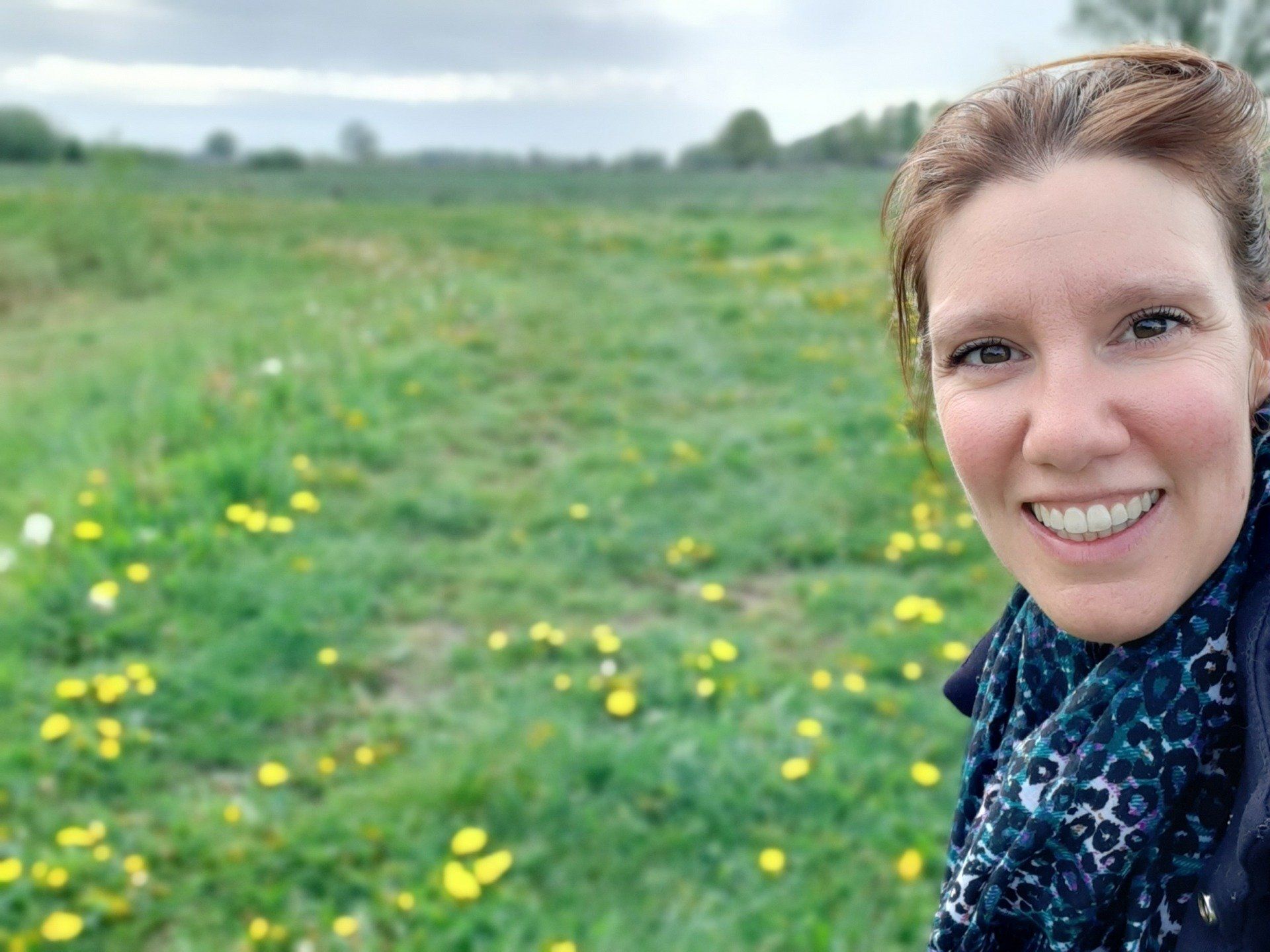 Joyce lachend in de buitenlucht, in een veld met groen gras en gele bloemen.