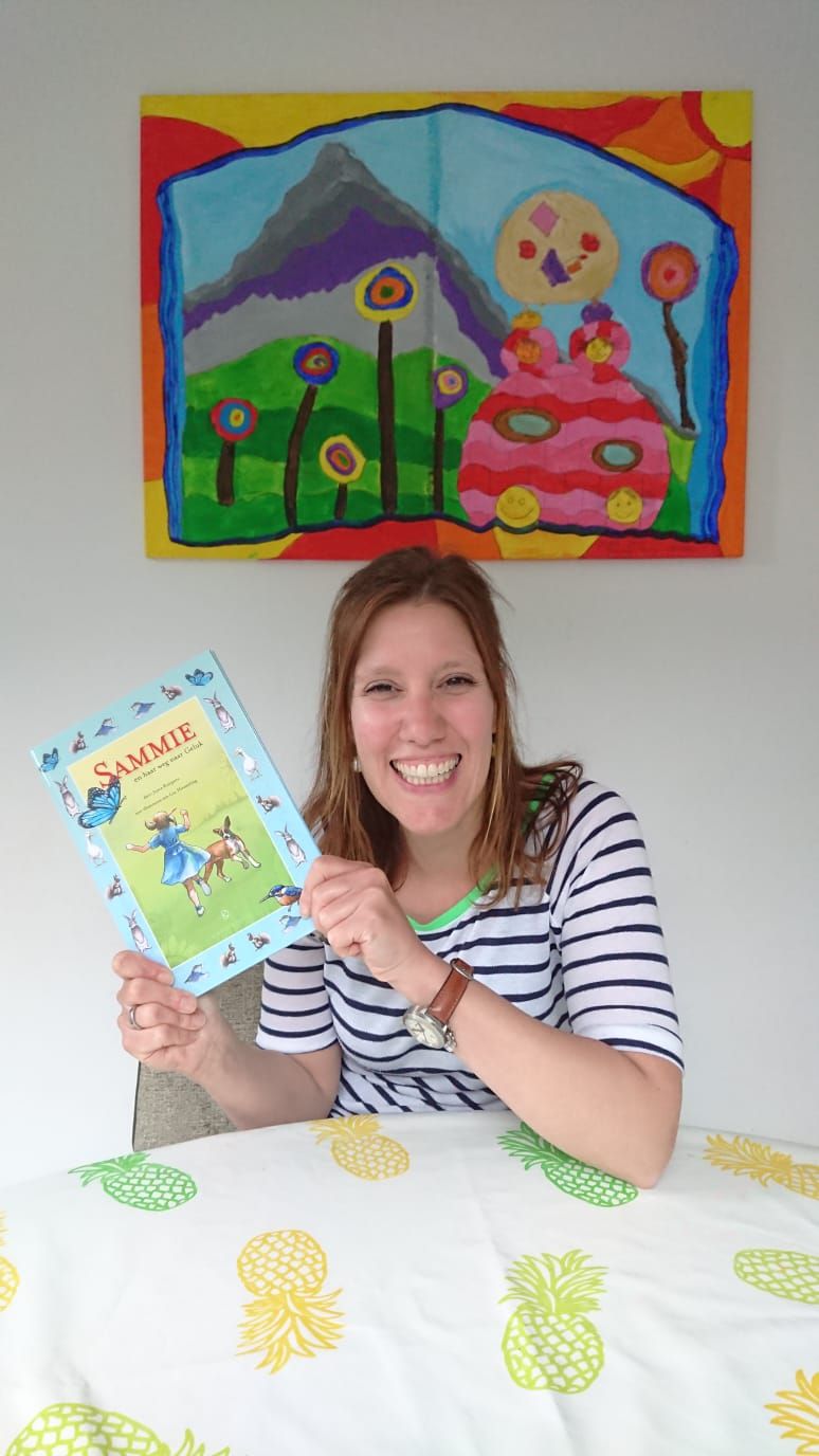 Een glimlachende vrouw zit aan een tafel met een boek in haar hand. Aan de muur hangt een kleurrijk kunstwerk.