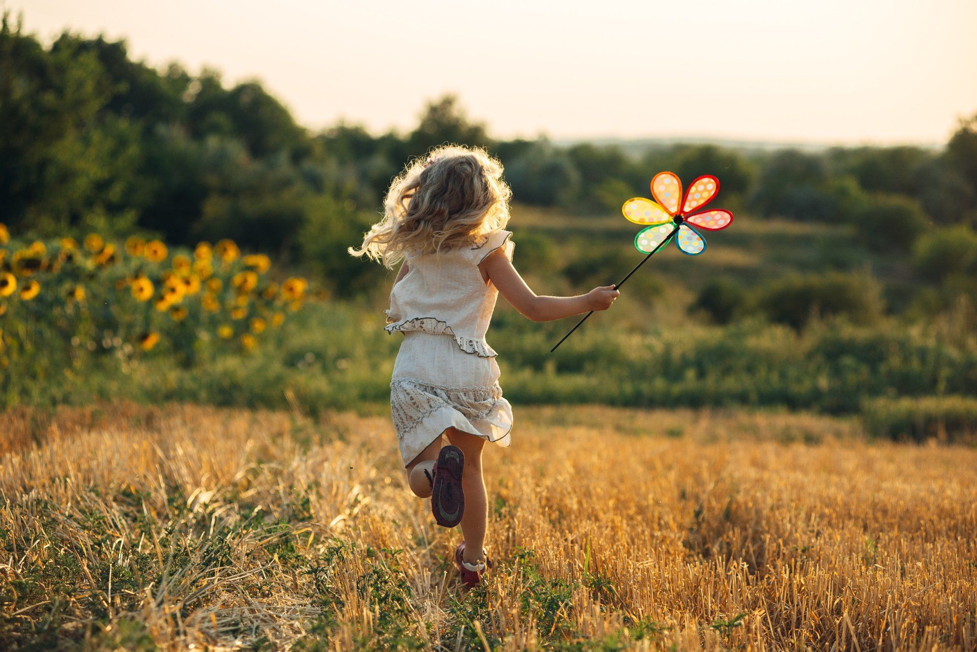 Een meisje rent door een veld met een kleurrijk windmolentje in haar hand. Zonnebloemen en bomen op de achtergrond.