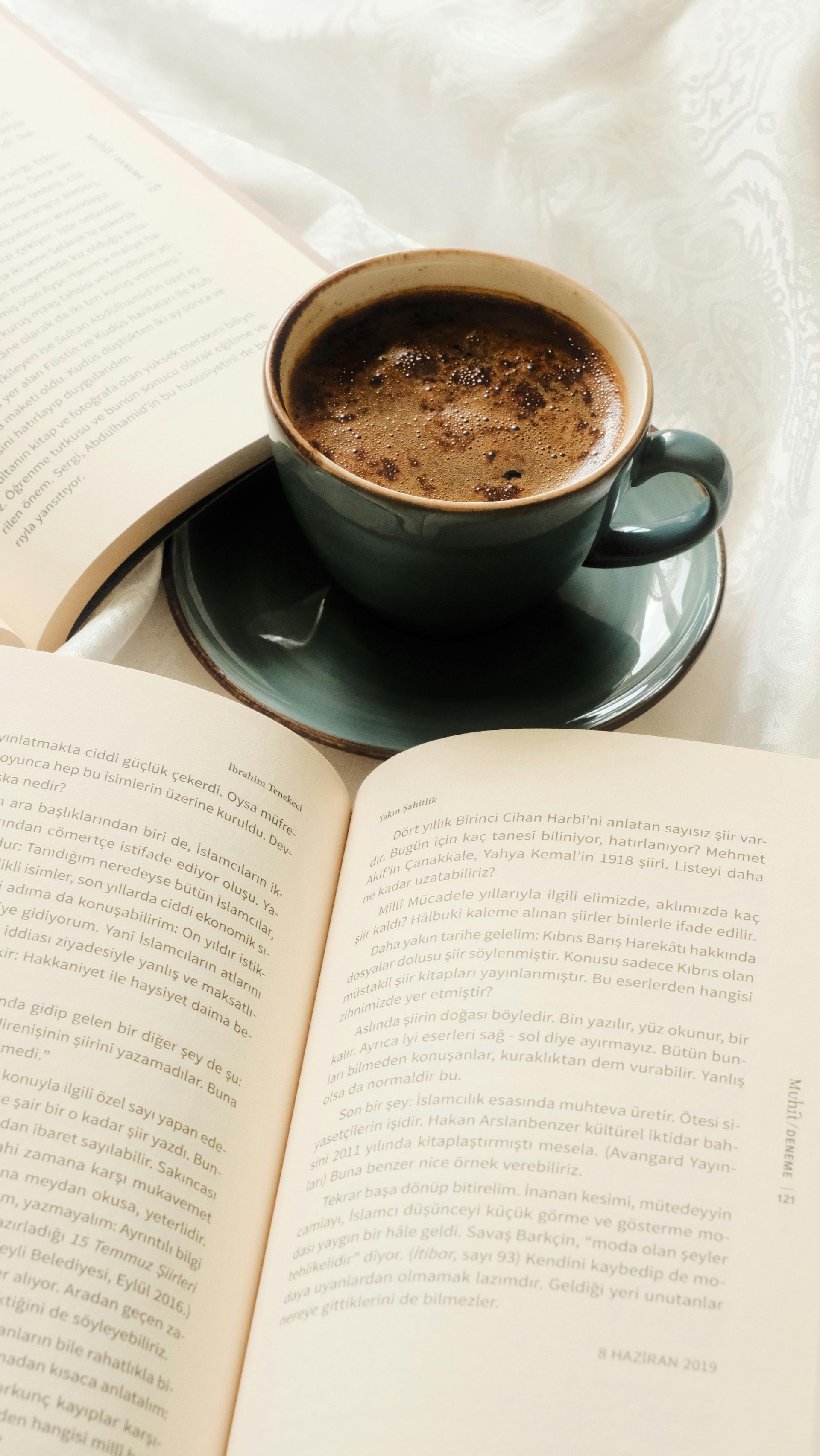 Een open boek naast een donkergroene kop koffie op een wit bed of tafel.