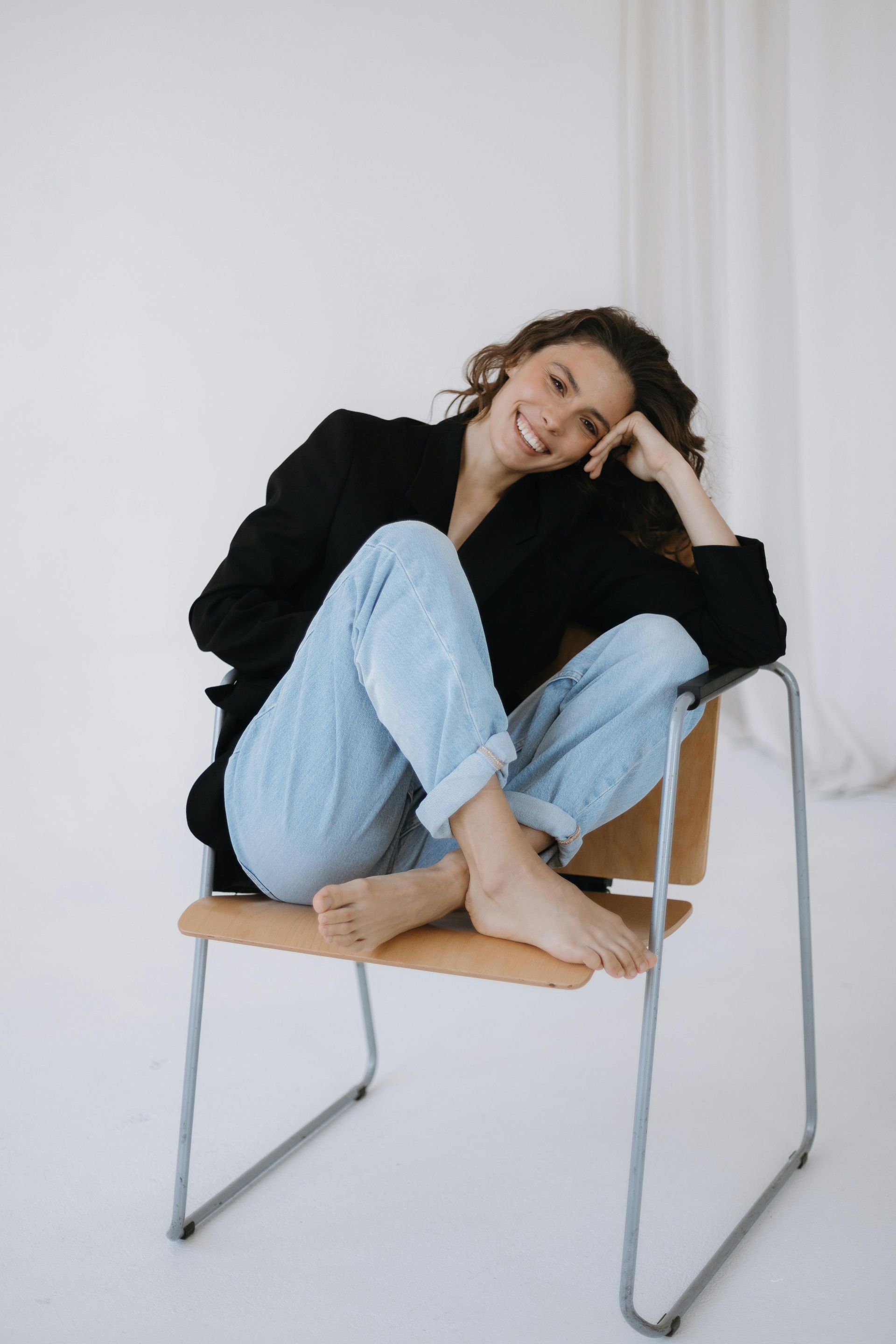 Een vrouw in een zwarte blazer en jeans zit glimlachend op een stoel binnenshuis.