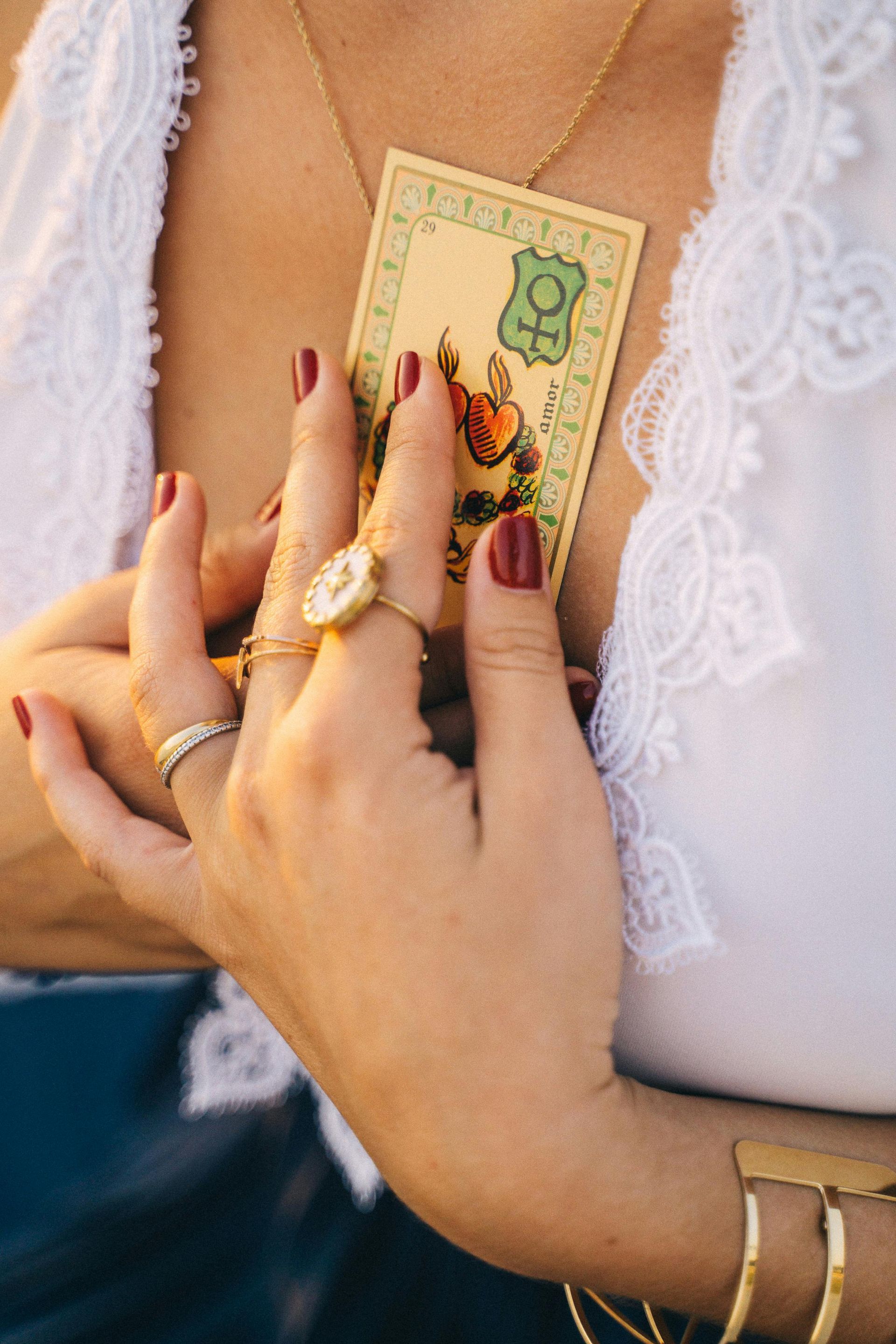Persoon met een tarotkaart met groene rand en gouden sieraden. Rode nagellak op de vingers. Wit kanten topje.