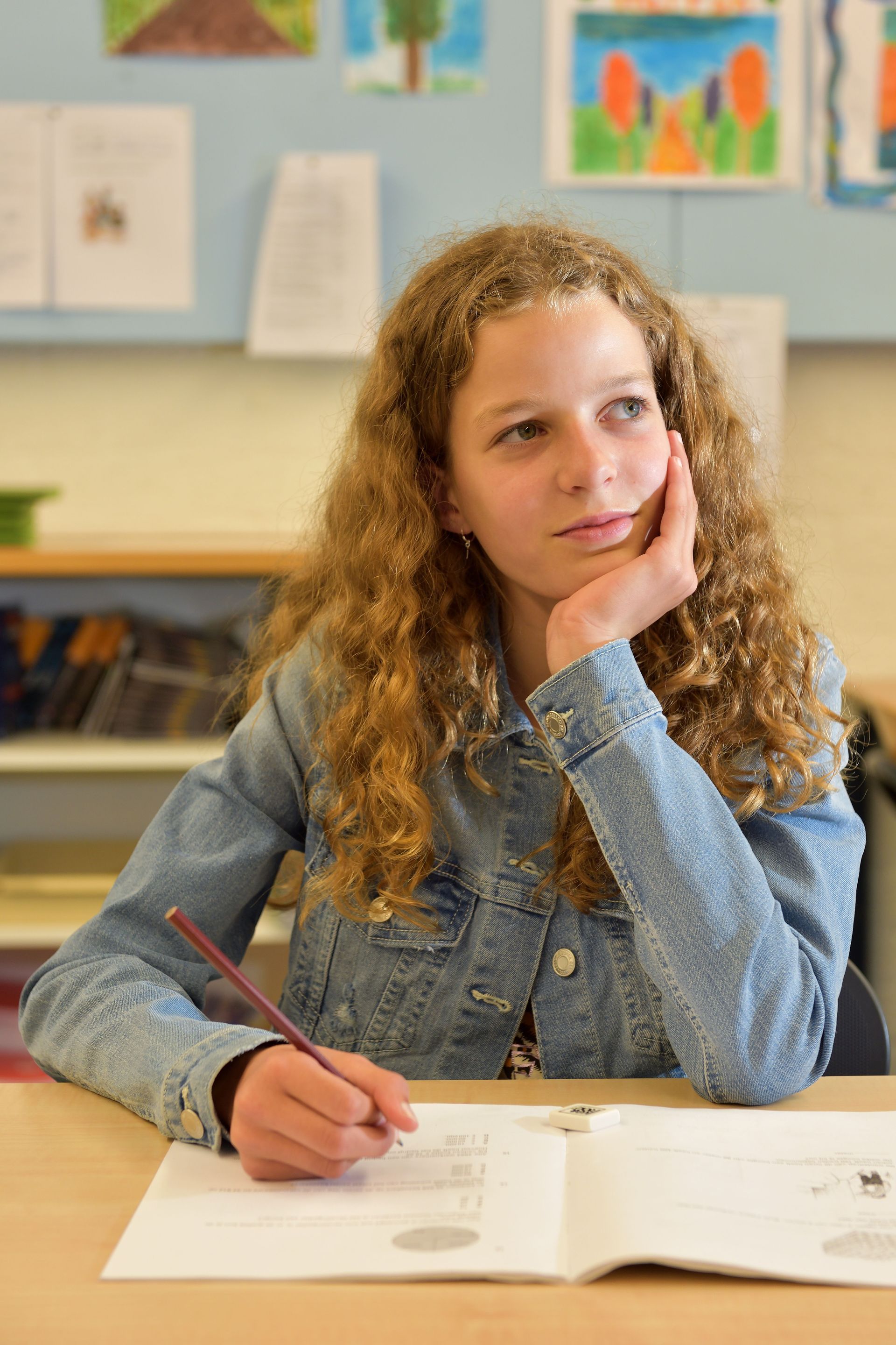 Student met krullend haar zit aan een bureau, schrijft in een open notitieboek en kijkt peinzend voor zich uit