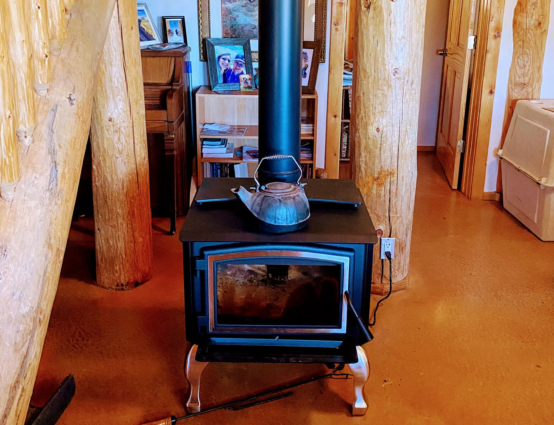A black wood-burning stove sits between two rough-hewn log pillars in a rustic room with orange-brown flooring.