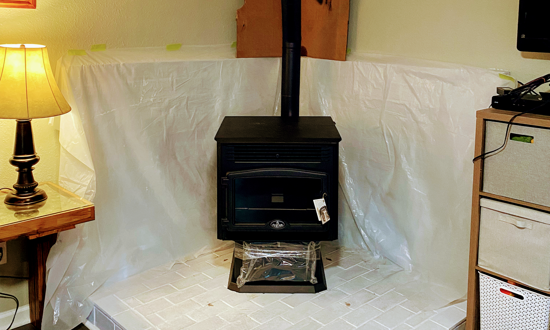 Black wood stove in a room covered with plastic sheeting, with a lit lamp on the left and a cabinet on the right