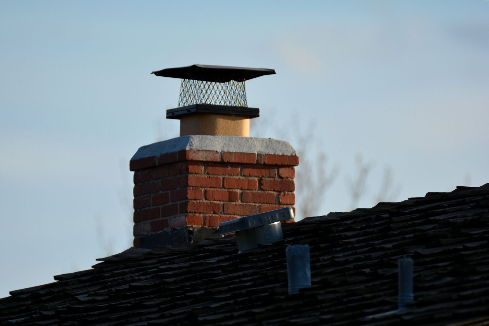 Brick chimney with a black cap rising above a dark shingled roof against a blue sky