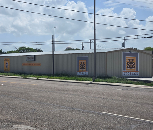 Storage facility with corrugated metal fencing and signs along a roadside under a cloudy sky
