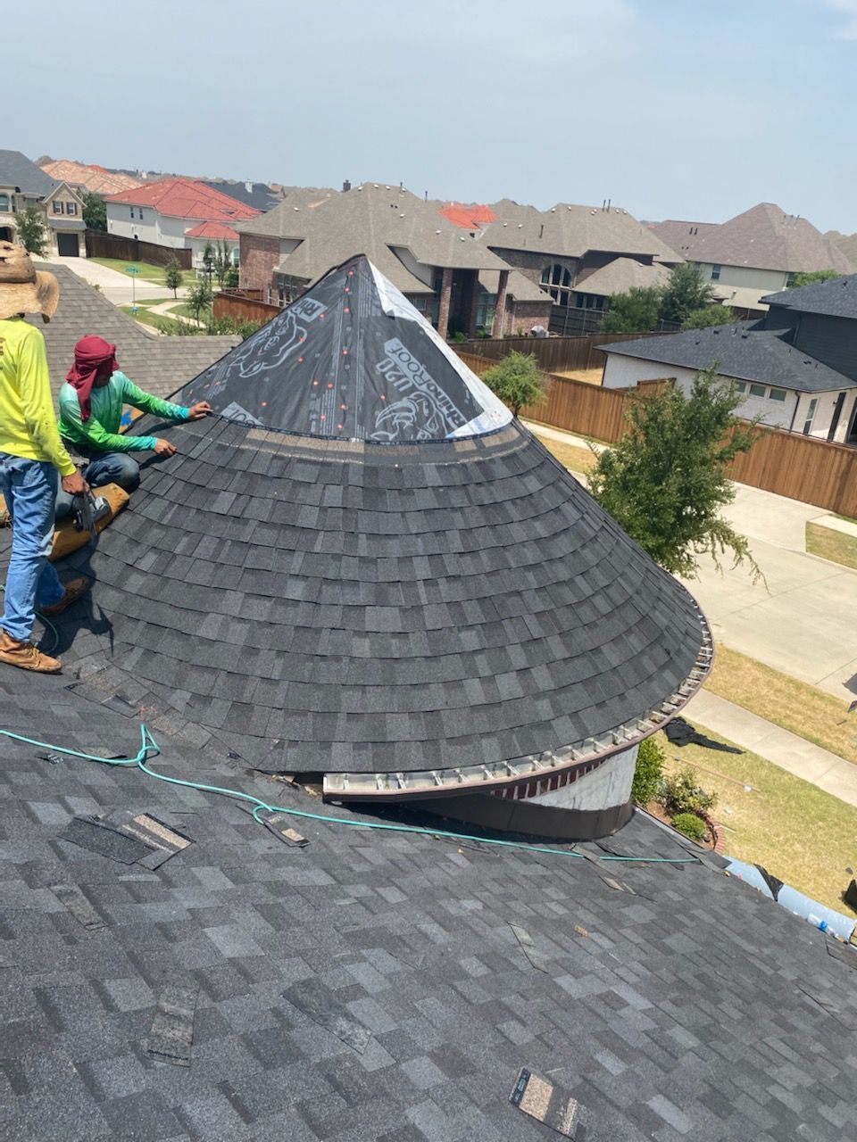 Two roofers installing shingles on a conical roof section, suburban houses in background.