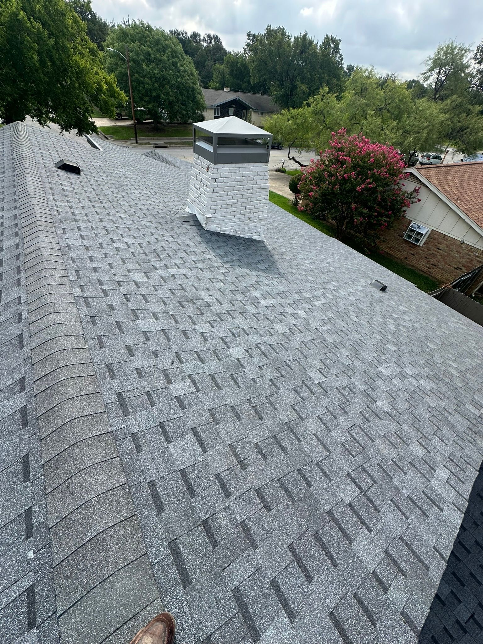 Gray asphalt shingle roof with chimney, trees in background, sunny day.
