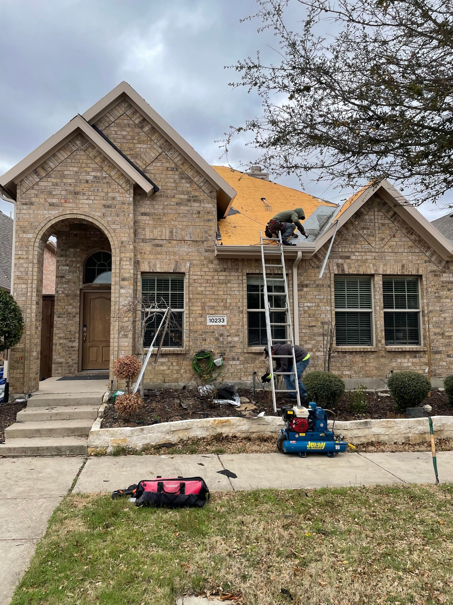 Roofers on ladder repairing roof of a brick house on a cloudy day. Tool bag and equipment sit on lawn.