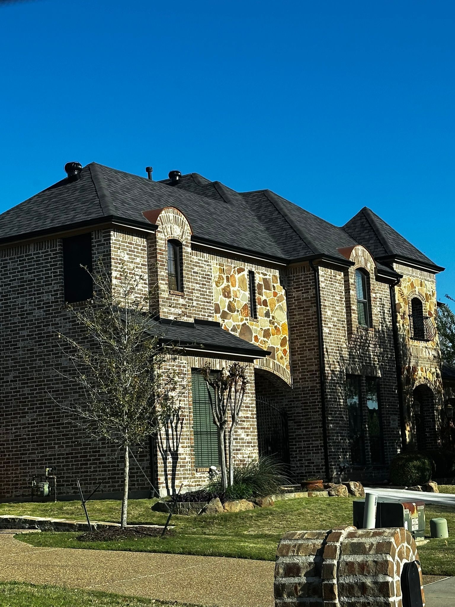Two-story brick and stone house under a bright blue sky; a mailbox is in the foreground.