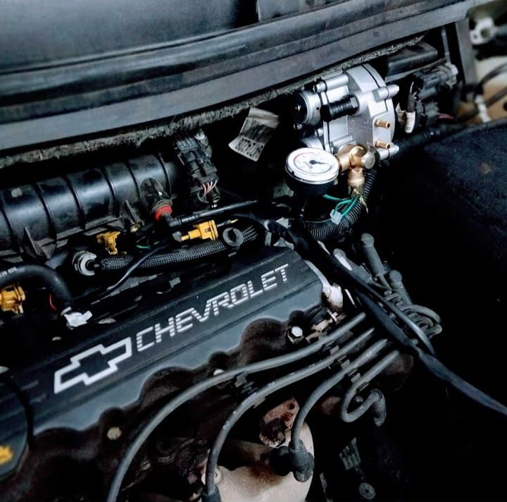 Compartimento del motor de un Chevrolet con tapa de válvulas negra y componentes del carburador visibles bajo el capó.