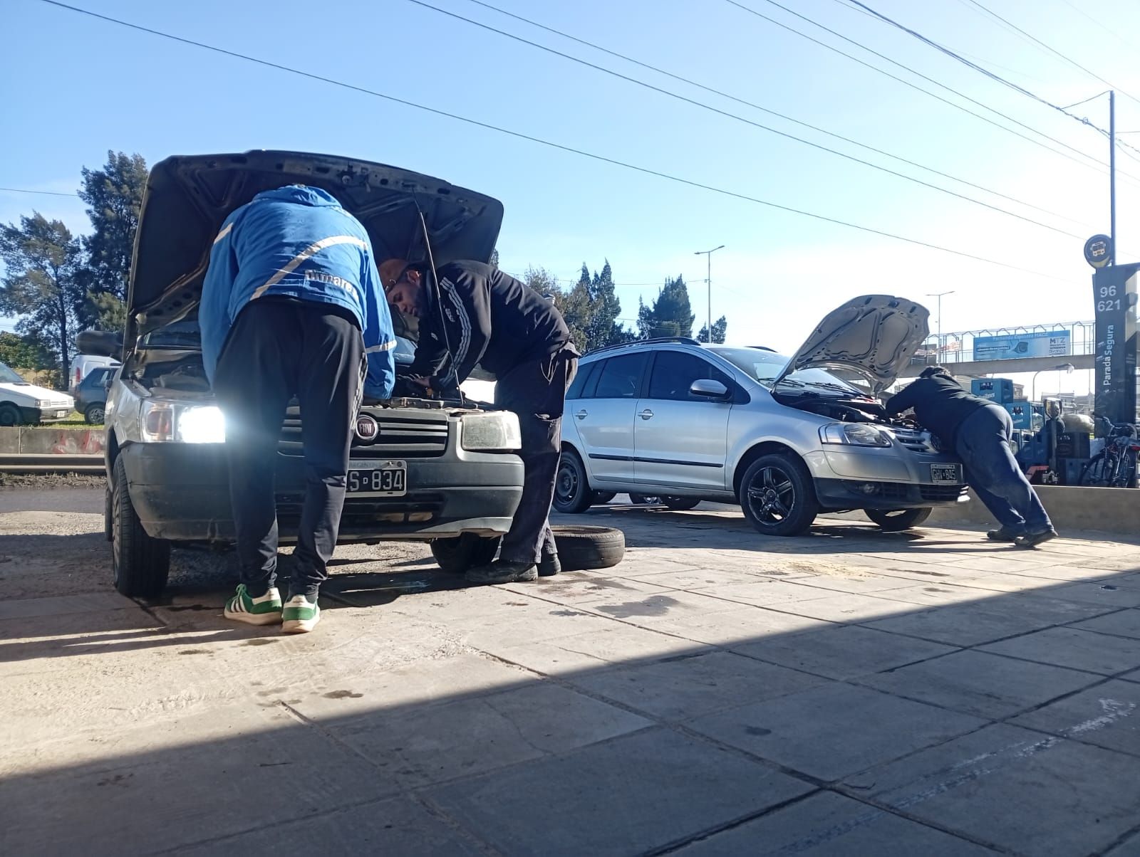 Varias personas trabajan debajo de dos coches con los capós abiertos en una acera soleada tras una avería en la carretera.