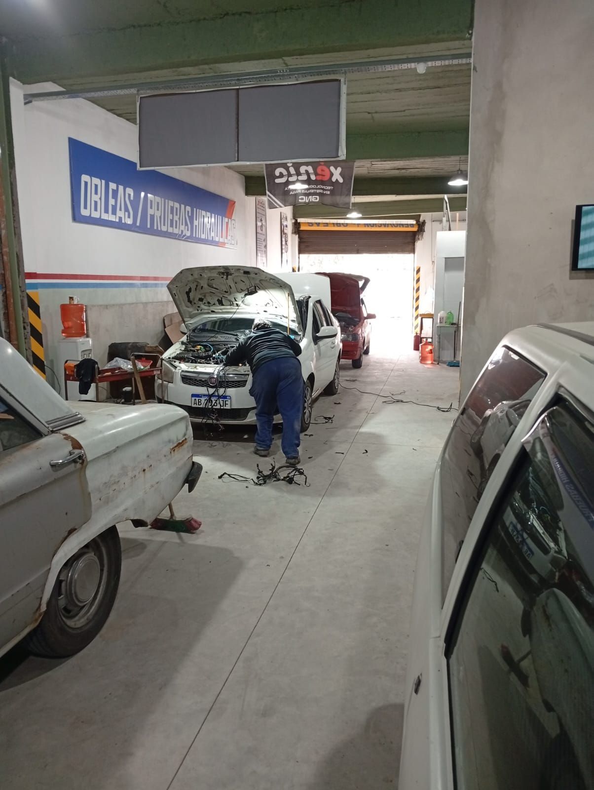 Un mecánico trabaja en un coche con el capó abierto en un estrecho pasillo de un garaje, rodeado de vehículos aparcados.