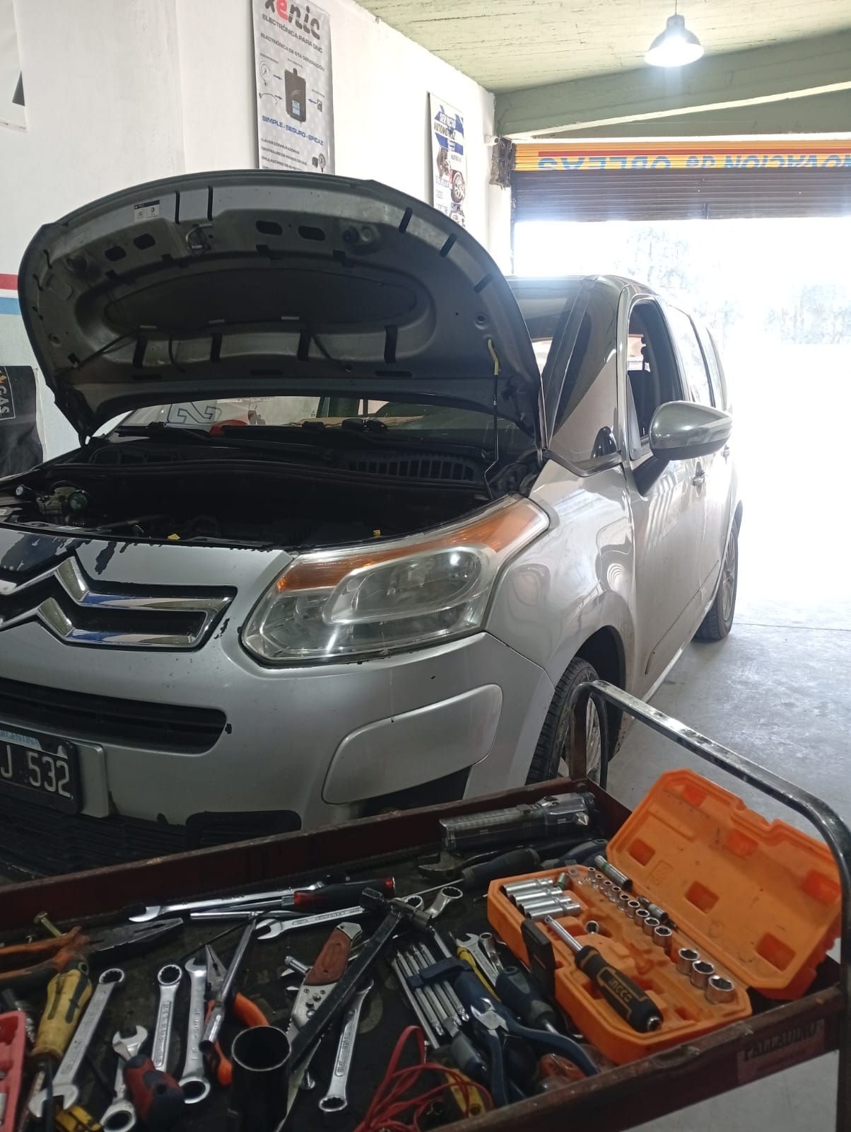 Un SUV plateado con el capó abierto en un garaje, con herramientas esparcidas sobre un banco de trabajo delante.