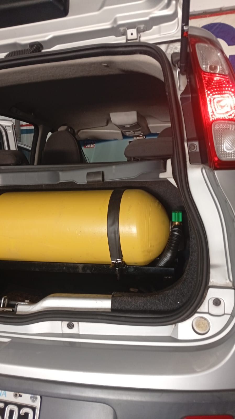 Depósito de gasolina amarillo en el maletero abierto de un coche plateado, sujeto con mangueras negras.