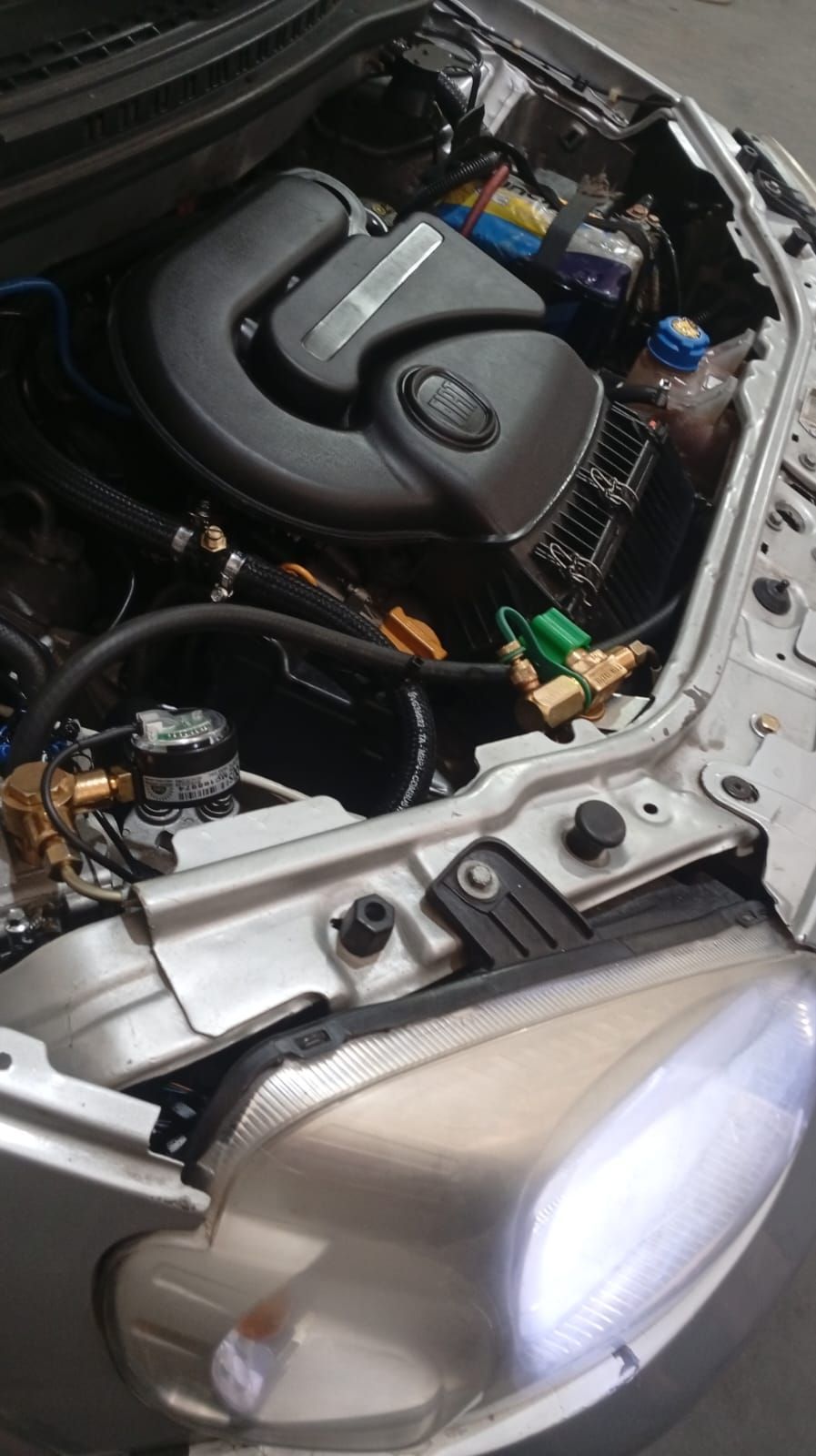 Compartimento del motor del coche con batería, mangueras y faro plateado en primer plano.