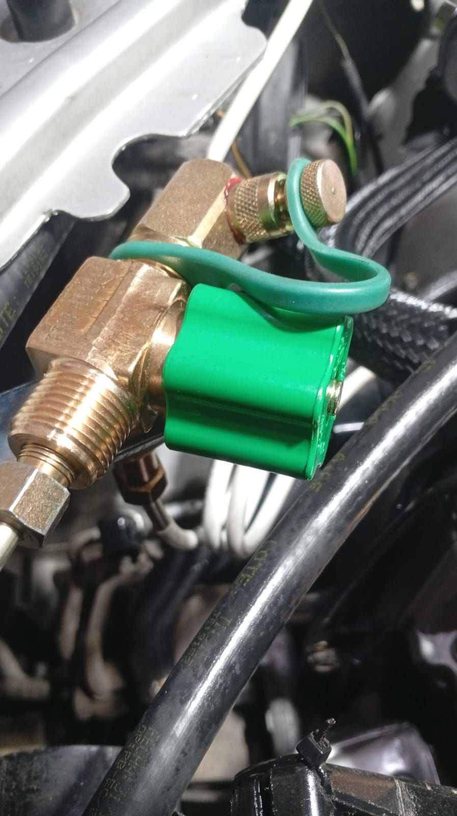 Conector eléctrico de latón con cables verdes sujeto a un componente del motor de un automóvil.