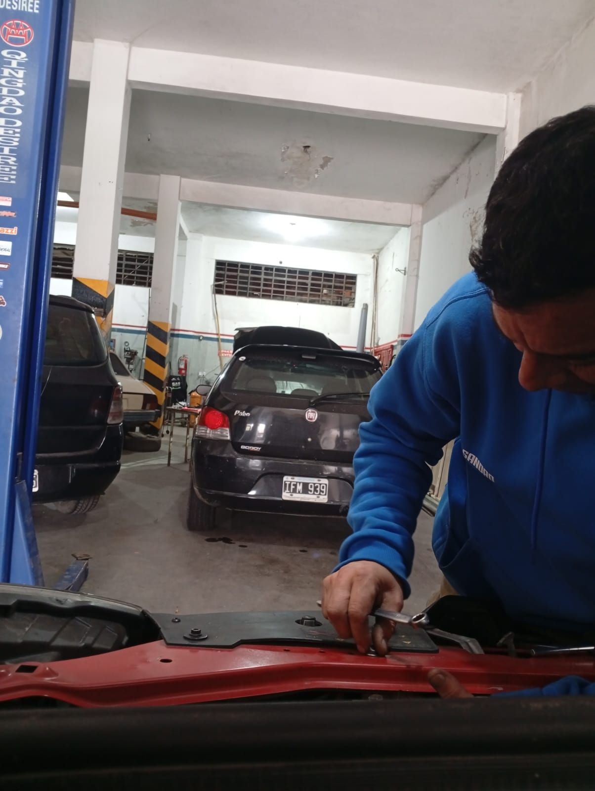 Mecánico con sudadera azul trabajando bajo el capó de un coche dentro de un taller mecánico.