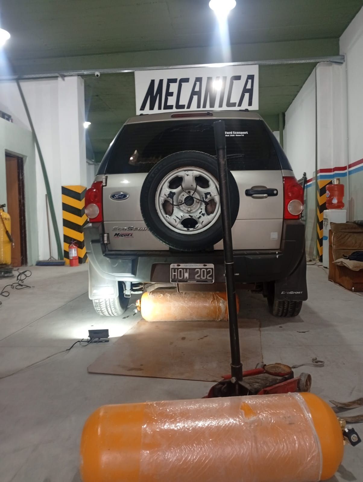 SUV plateado en un elevador dentro de un taller mecánico, con un gato hidráulico y un tanque naranja en primer plano.