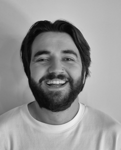 Een zwart-witportret van een lachend persoon met een baard, gekleed in een T-shirt met ronde hals, tegen een effen achtergrond.
