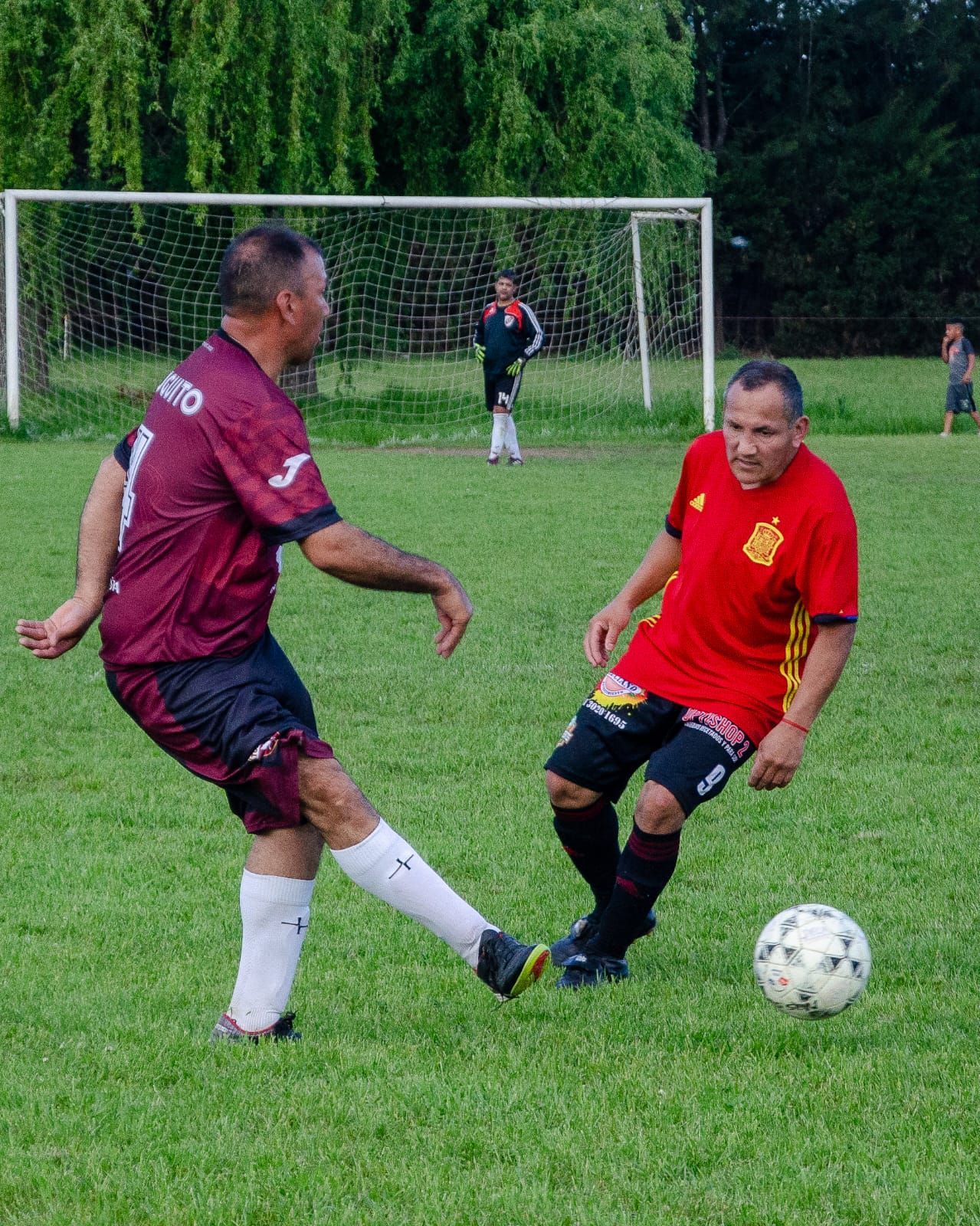 Dos jugadores uniformados de fútbol compiten por un balón en un campo de césped frente a una red de portería.