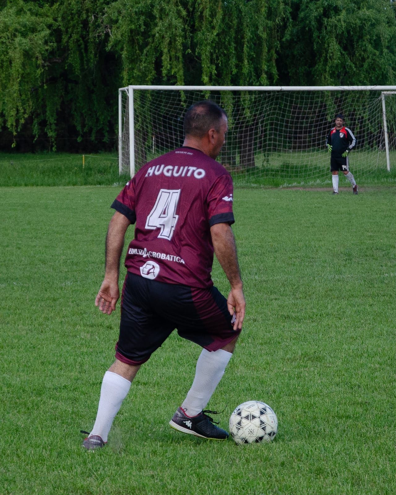 Un jugador de fútbol con una camiseta marrón con el número 4 dribla un balón en un campo verde cerca de una portería