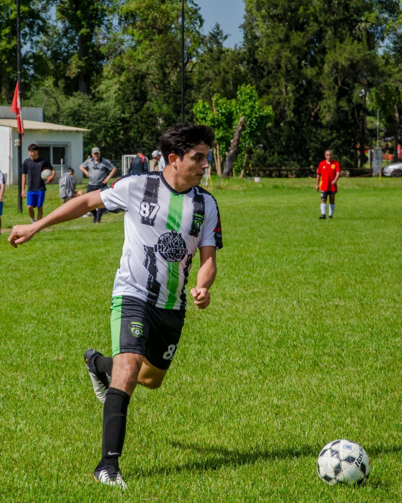 Un jugador con un uniforme de fútbol verde, negro y blanco dribla un balón de fútbol en un campo de césped soleado.
