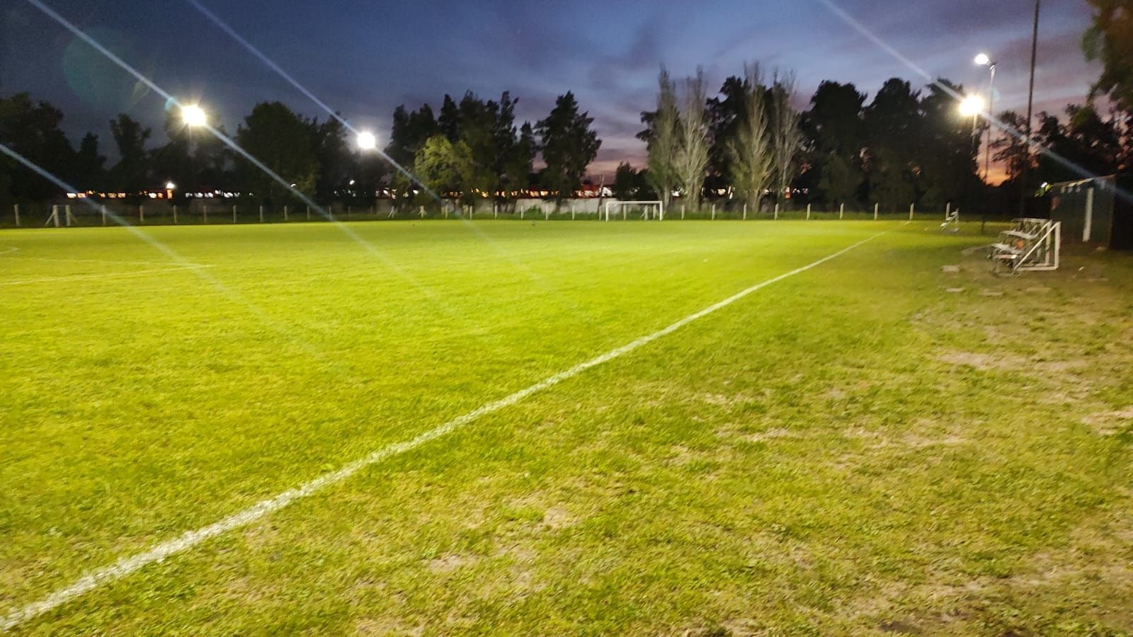 Un campo de fútbol de césped iluminado al anochecer, con líneas blancas de límite, luces de estadio y postes de gol.