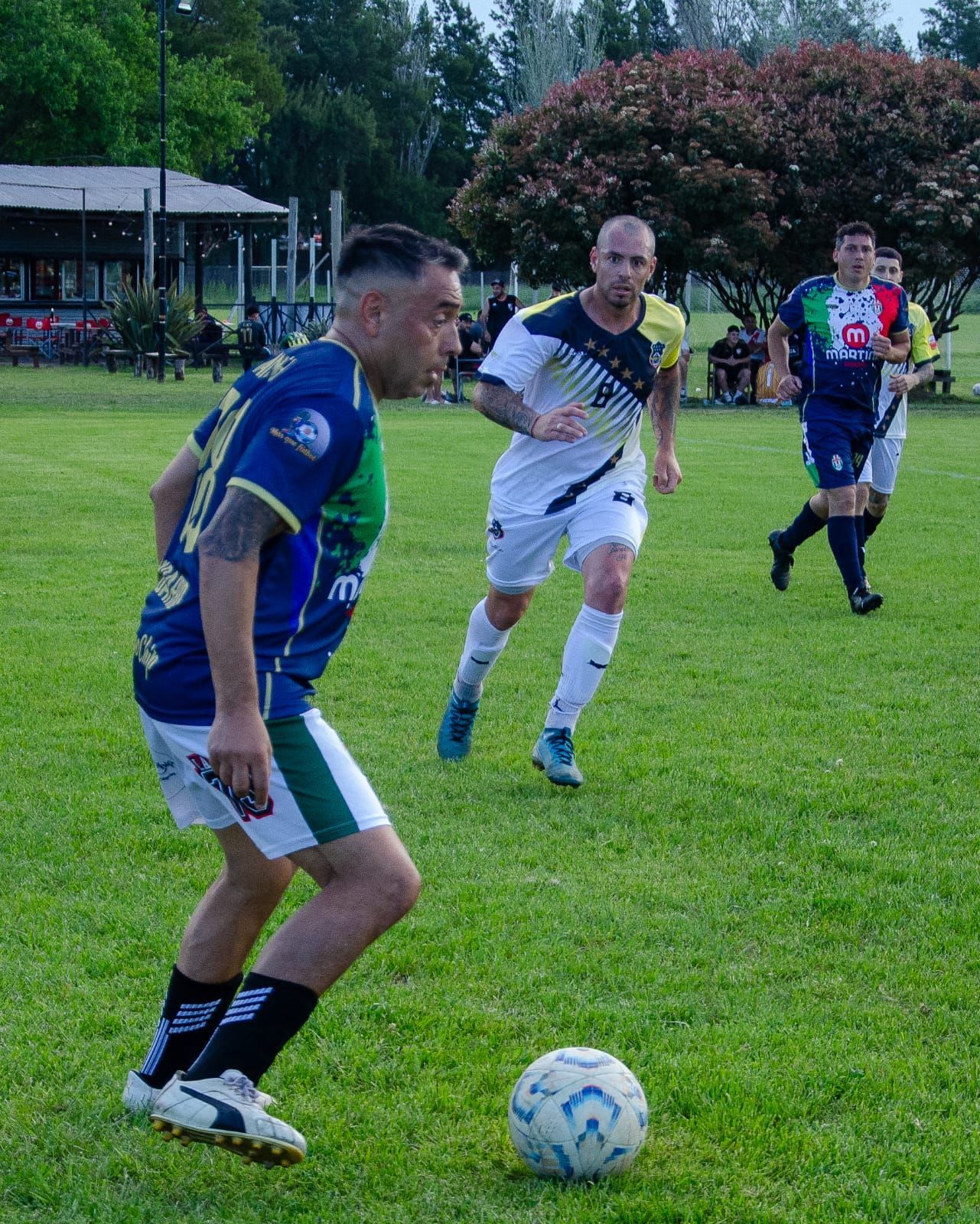 Un jugador con uniforme azul y verde dribla un balón de fútbol en un campo de césped mientras otros corren hacia él.