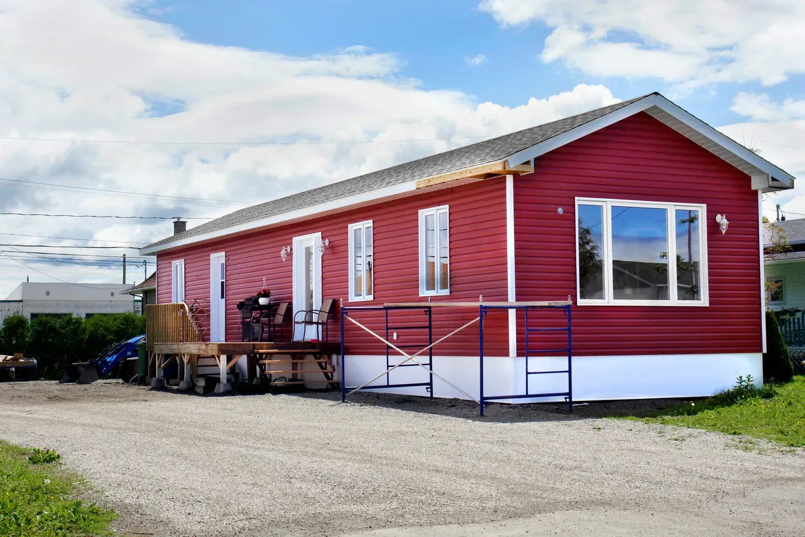 A red manufactured home with white trim and a front deck, set on a gravel lot under a blue sky with clouds.