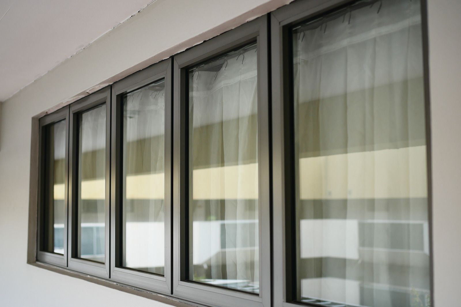 A row of four side-by-side rectangular windows with dark grey frames and light, sheer curtains visible inside.