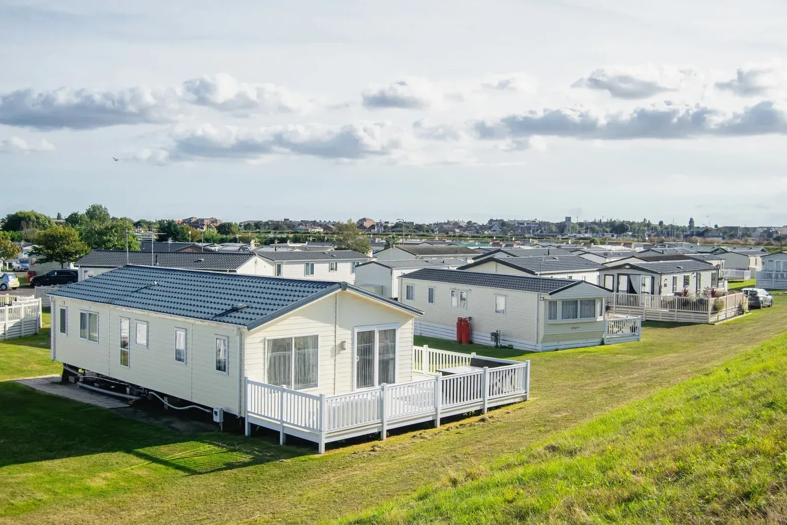 A row of light-colored mobile homes with white decks arranged on a grassy field under a cloudy blue sky.